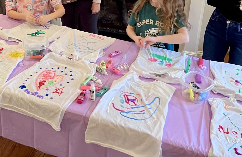 Decorated white t-shirts on a table with puffy paint pens during a kids birthday party activity