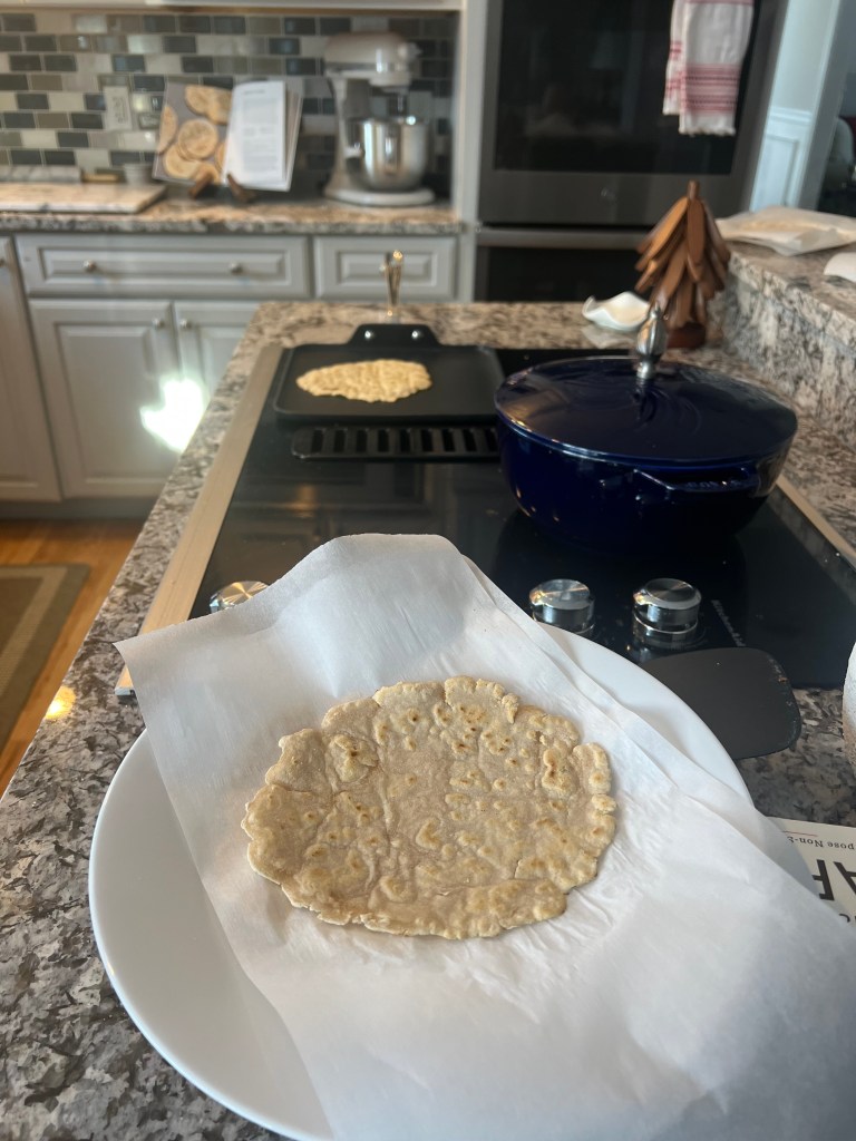 Cooked cassava flour tortilla with another tortilla cooking in a skillet, with the Downshiftology cookbook open to the recipe in the background.