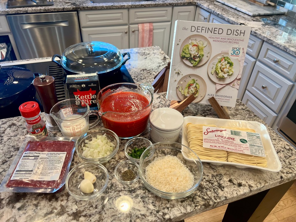 The Defined Dish cookbook with ingredients for weeknight bolognese on a kitchen counter