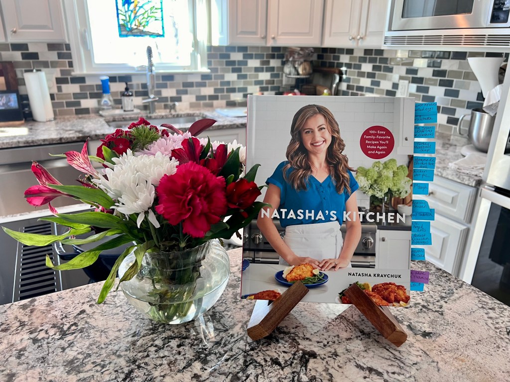 A copy of Natasha's Kitchen cookbook on the kitchen counter with flowers in a vase.