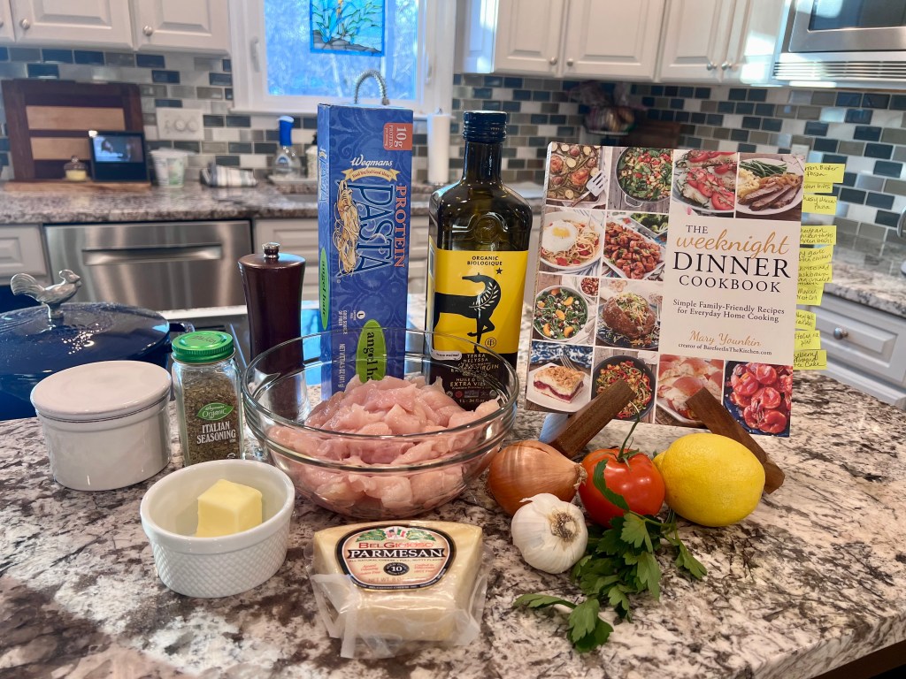 The Weeknight Dinner cookbook on the counter with ingredients for garlic butter pasta and Italian chicken bites arranged beside it.