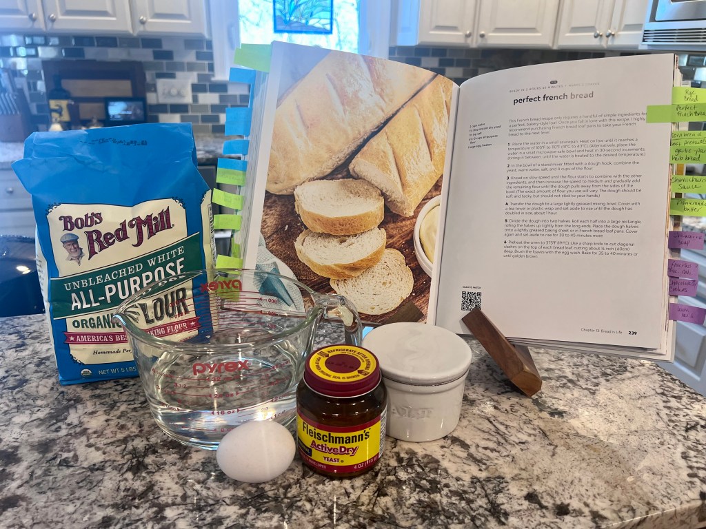 Cookbook open to a homemade French bread recipe with measured ingredients on the kitchen counter.