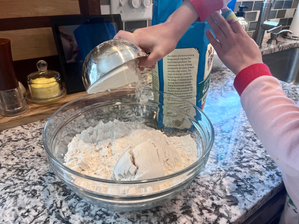 Child’s hands pouring flour into a mixing bowl while helping make homemade French bread from scratch.