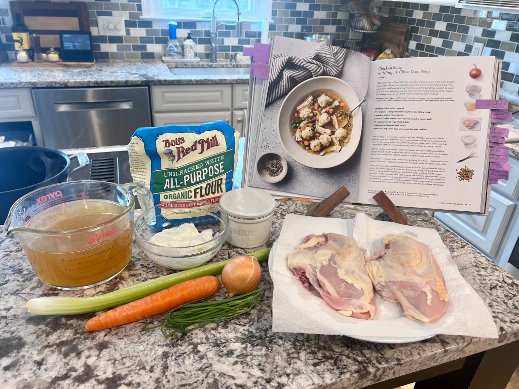 Open cookbook on a kitchen counter displaying a chicken soup with yogurt dumplings recipe, surrounded by fresh ingredients including carrots, celery, flour, yogurt, and herbs.
