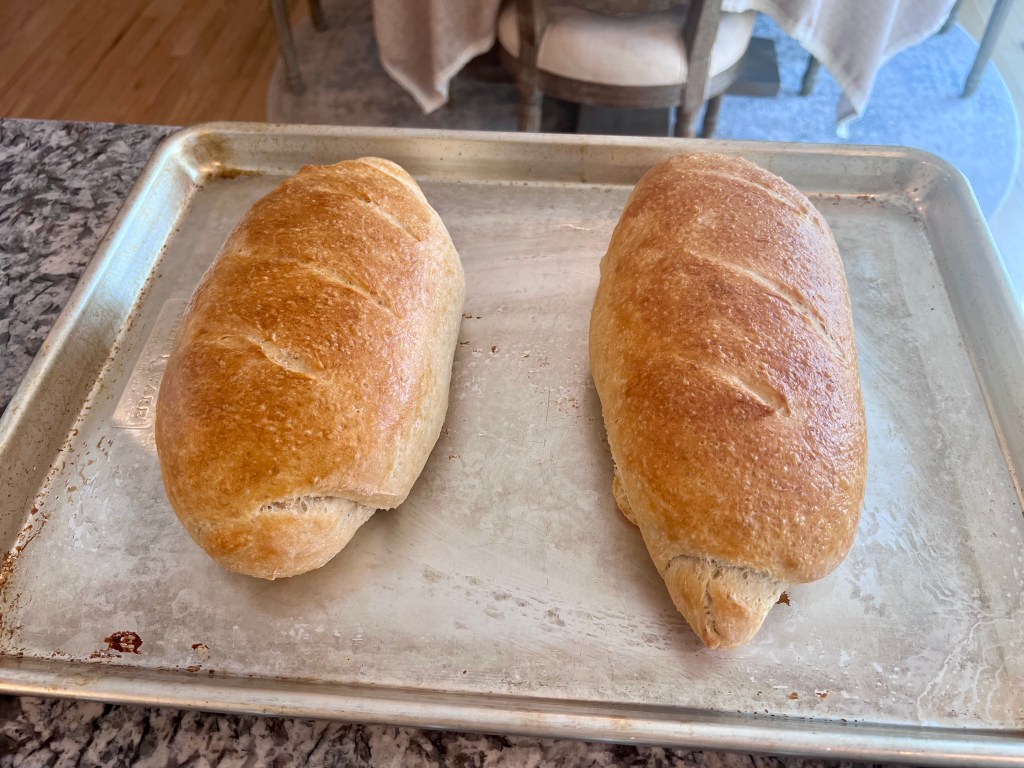Two golden brown homemade French bread loaves fresh out of the oven on a baking sheet.