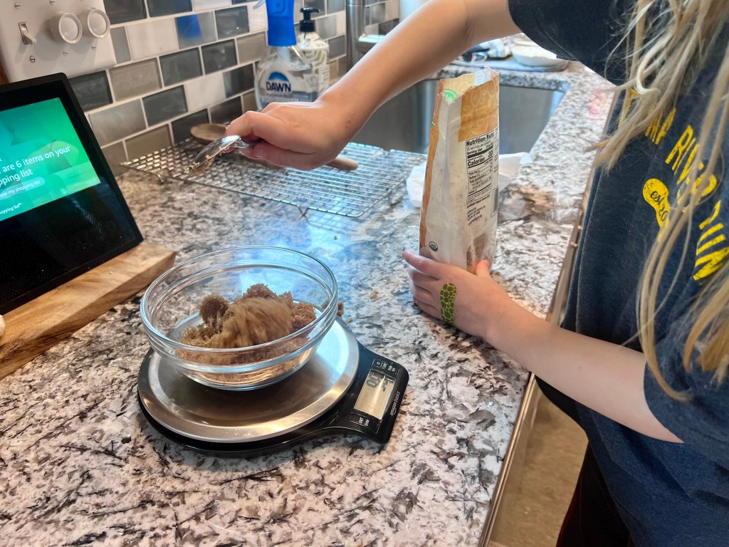 Child measuring brown sugar on a kitchen scale while helping bake homemade banana bread