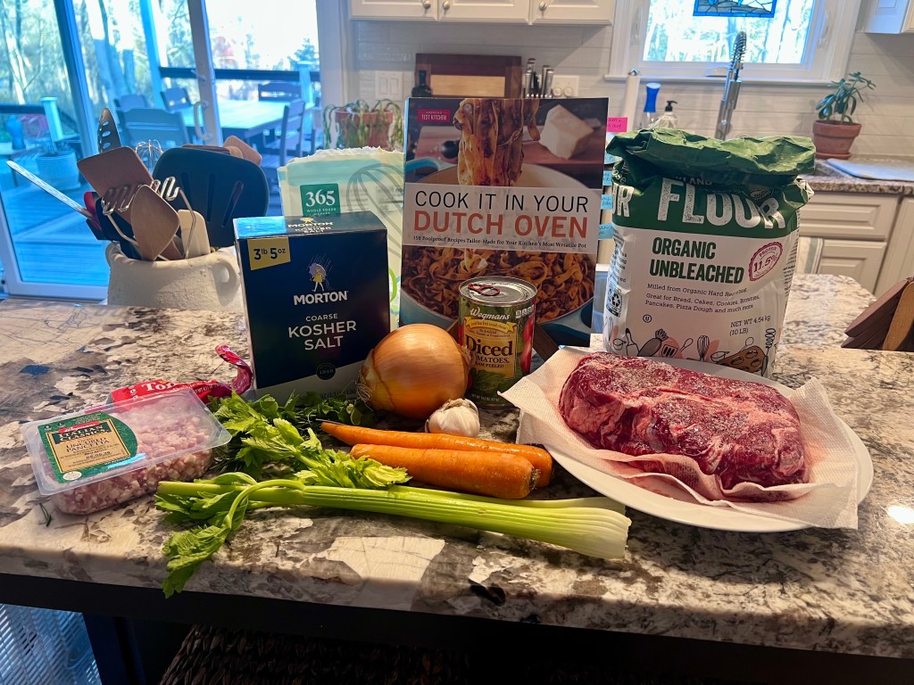 Barolo braised beef ingredients with chuck roast, vegetables, and red wine next to Cook It in Your Dutch Oven cookbook on kitchen counter