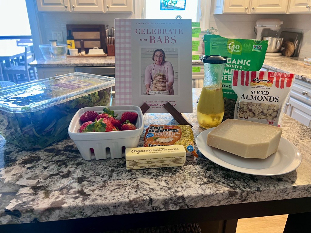 Strawberry salad ingredients on counter with fresh strawberries, greens, ramen noodles, almonds, and cookbook