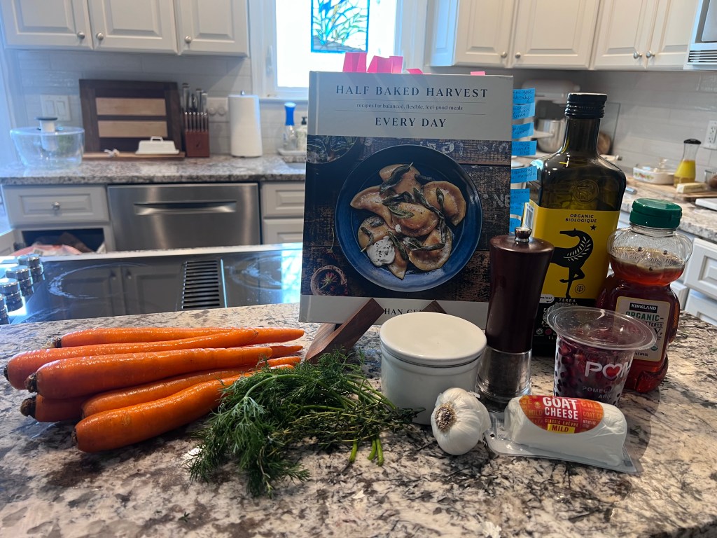 Ingredients for honey roasted carrots on the counter with carrots, herbs, goat cheese, pomegranate, and the Half Baked Harvest Every Day cookbook